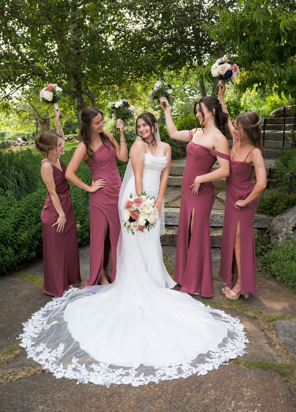 Wedding party in burgundy dresses posing outdoors with bouquets against green foliage.