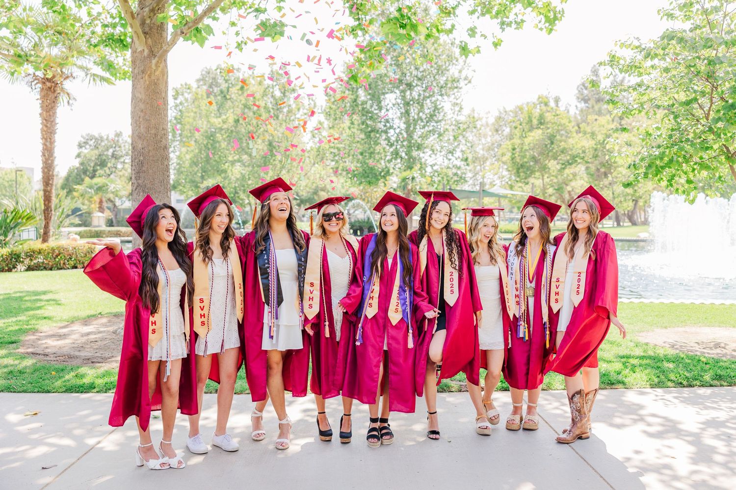 Group of graduates in bright red caps and gowns pose outdoors in a sunny park setting with trees and greenery.
