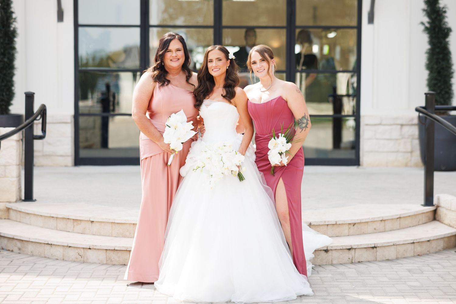 A bridal party poses together at Bella Cavalli's entrance, wearing pink and burgundy dresses with white floral bouquets.