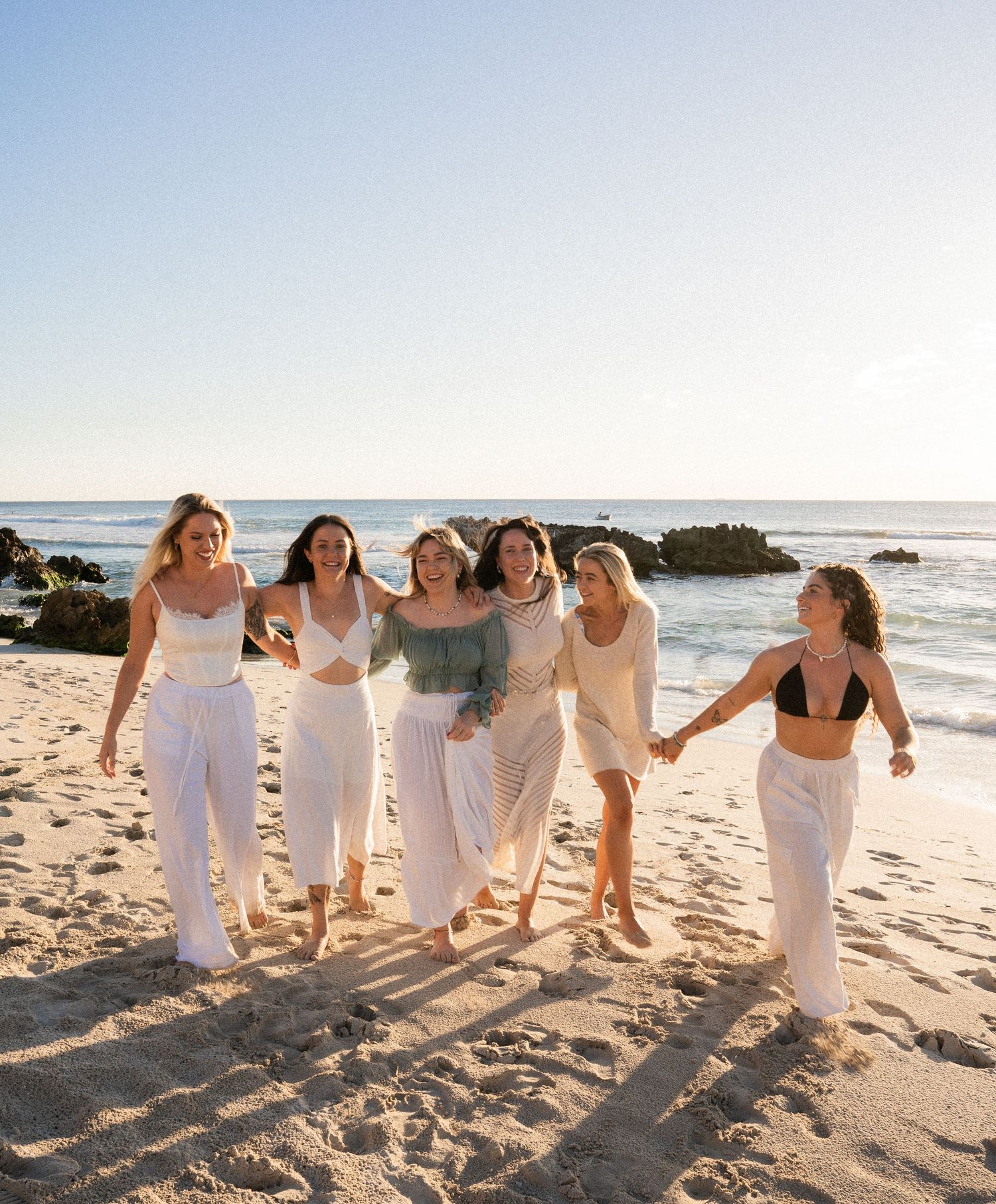 Group of wedding party attendants in white dresses walking and laughing together on a sunny beach at sunset.