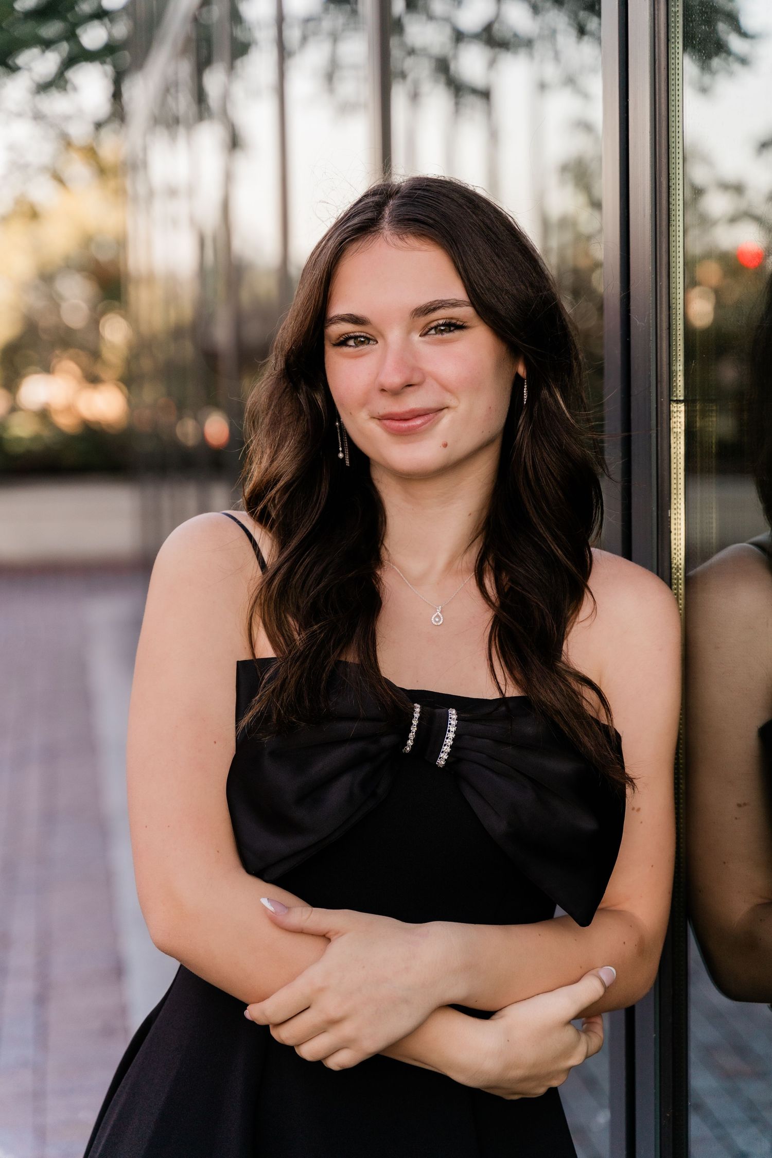 A high school senior in a black dress stands against a glass wall outdoors with trees visible in the background in Downtown Toledo session.