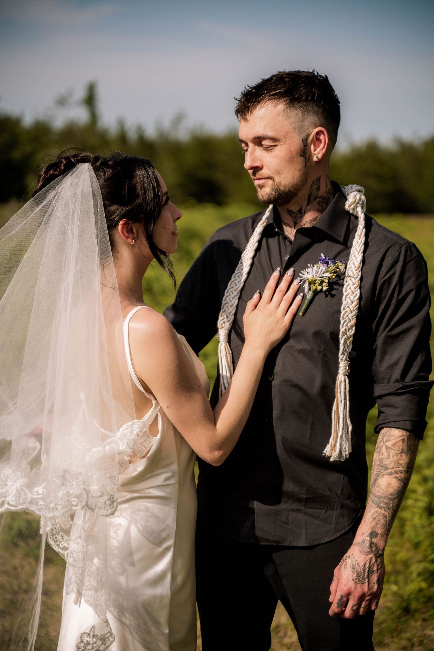 A bride in a white dress and veil touches the groom's chest tenderly during an outdoor wedding ceremony.