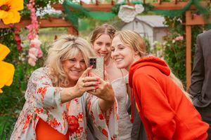 Three friends taking a selfie together among colorful garden flowers at an outdoor event.