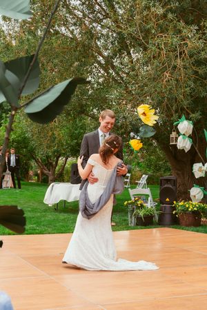 A wedding couple shares a joyful dance together on an outdoor wooden dance floor surrounded by trees and paper lanterns.