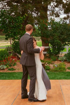 A couple shares their first dance together in formal attire on an outdoor wooden deck surrounded by lush greenery.