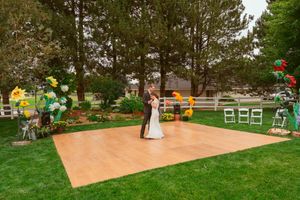 A rustic outdoor wedding ceremony setup with a wooden dance floor, garden decorations and white chairs.