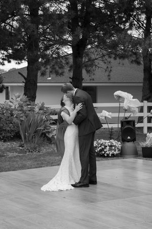 Black and white photo of newlyweds dancing together on an outdoor patio with pine trees in the background.
