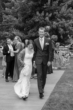A black and white photo of newlyweds walking down an outdoor aisle after their wedding ceremony.