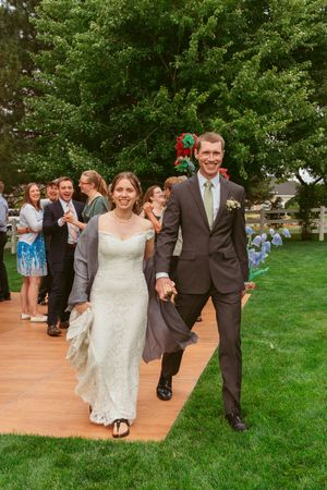 A couple in wedding attire walk together on a wooden pathway surrounded by guests and greenery at an outdoor ceremony.