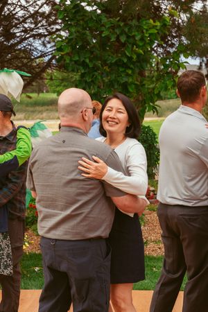 A group in coordinated grey outfits embrace in a celebratory group hug at an outdoor wedding venue.