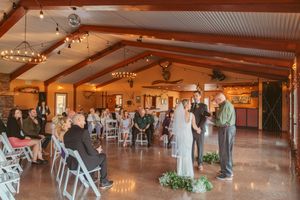 A rustic indoor wedding ceremony takes place in a wooden lodge venue decorated with string lights and natural accents.