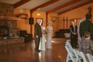 An indoor wedding ceremony in a rustic venue with exposed wooden beams and a stone fireplace.