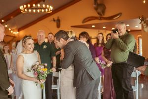 A couple shares an intimate moment during a wedding ceremony in a rustic venue decorated with string lights.