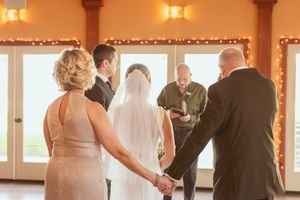 An intimate indoor wedding ceremony takes place under warm lighting with decorative garlands along the windows.