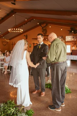 A wedding ceremony in progress inside a rustic venue with warm lighting and decorative greenery.