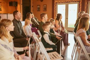 Wedding guests seated in white chairs watching an indoor ceremony in a warmly lit room.