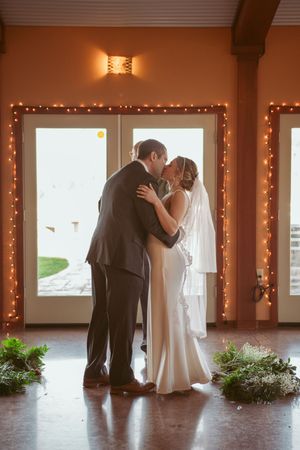 A romantic first kiss between newlyweds framed by fairy lights and large windows.