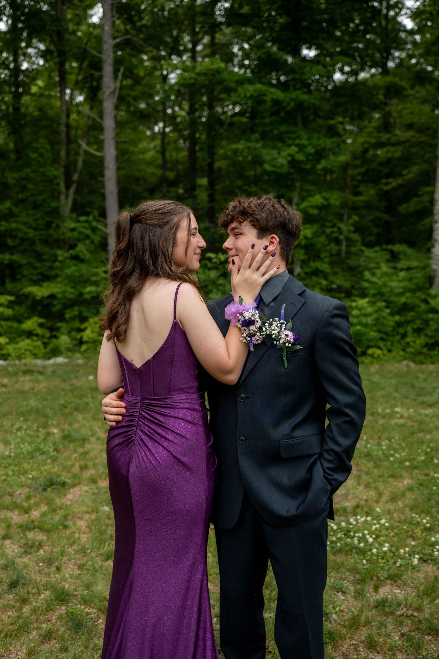 A person in a purple dress embraces someone in a navy suit outdoors with trees and wildflowers in the background.