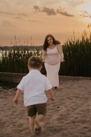 Young child in white shirt runs playfully toward a person in a white dress on a beach at sunset.