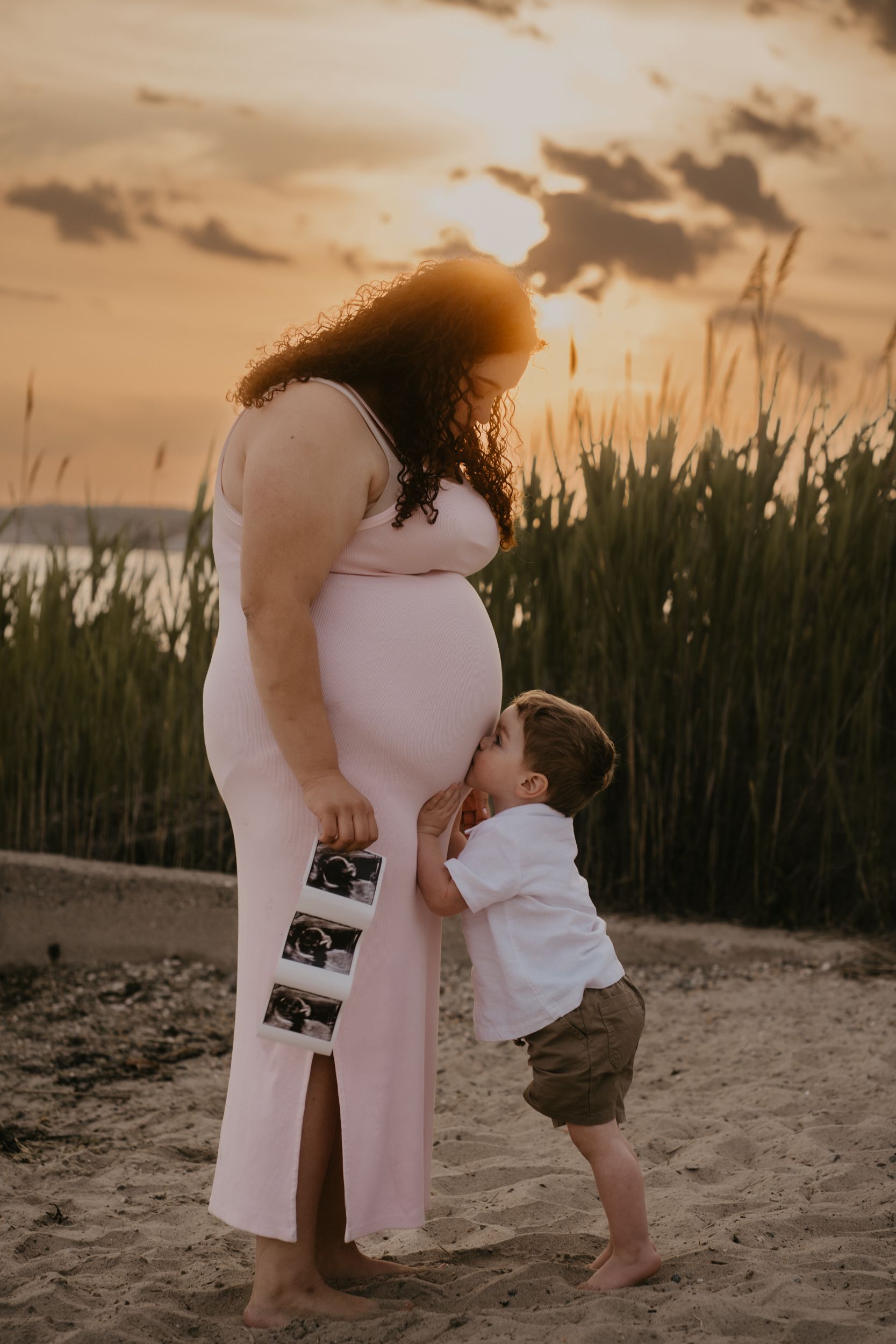 A pregnancy announcement photoshoot at sunset shows a toddler kissing a growing baby bump on the beach in Connecticut.