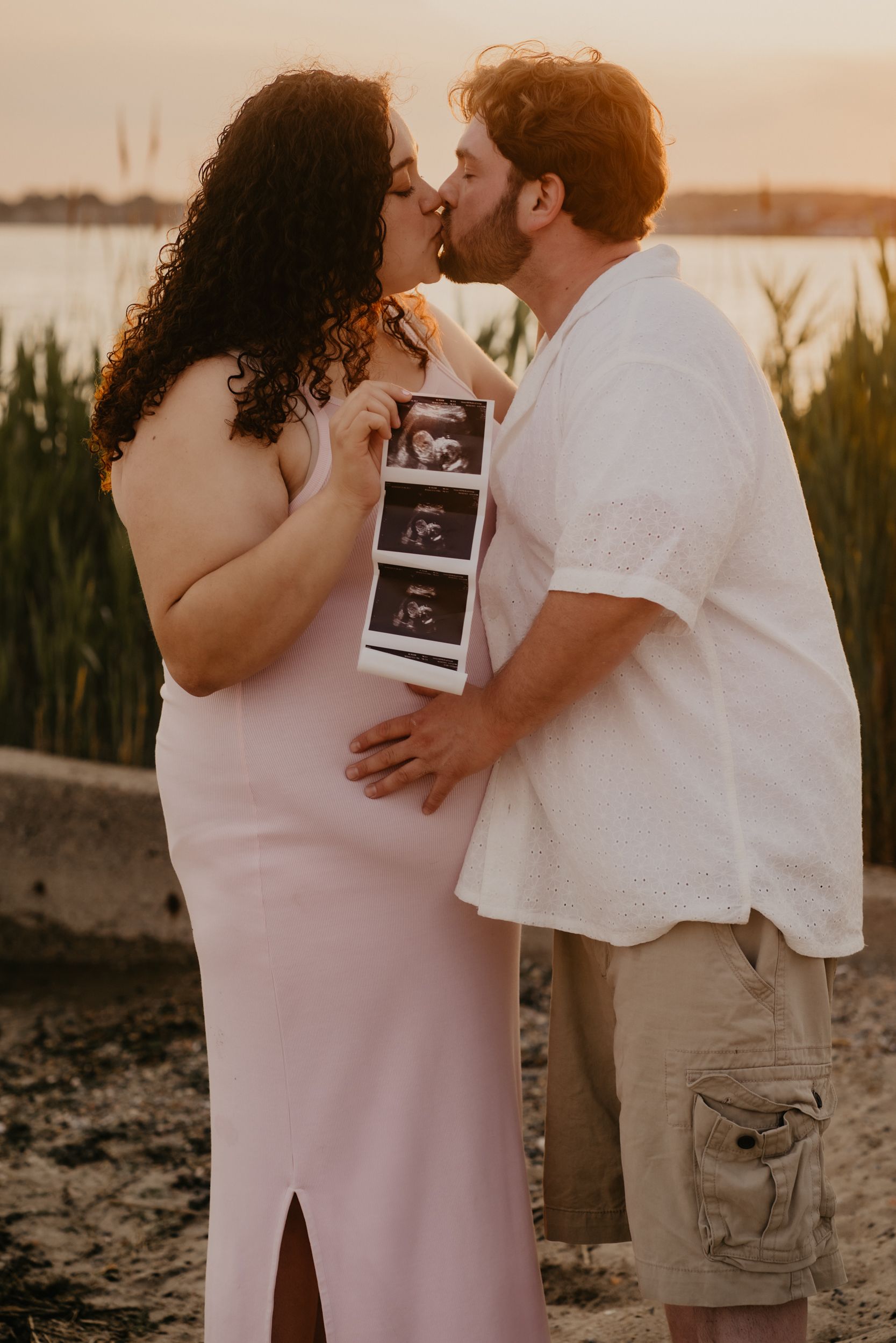 Romantic sunset maternity photoshoot featuring couple embracing while holding ultrasound photos on beach.