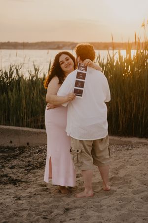 A couple shares a tender moment on the beach at sunset while holding an ultrasound photo for their pregnancy announcement.