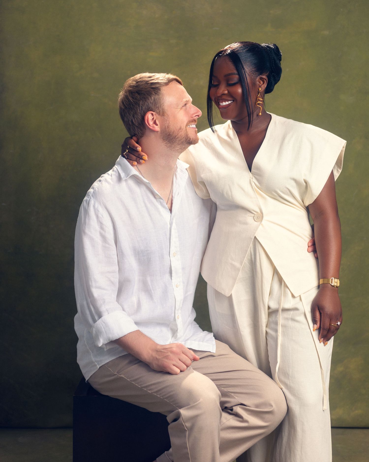 Two people in white clothing sit close together holding hands with one wearing a gold watch and white wrap dress.