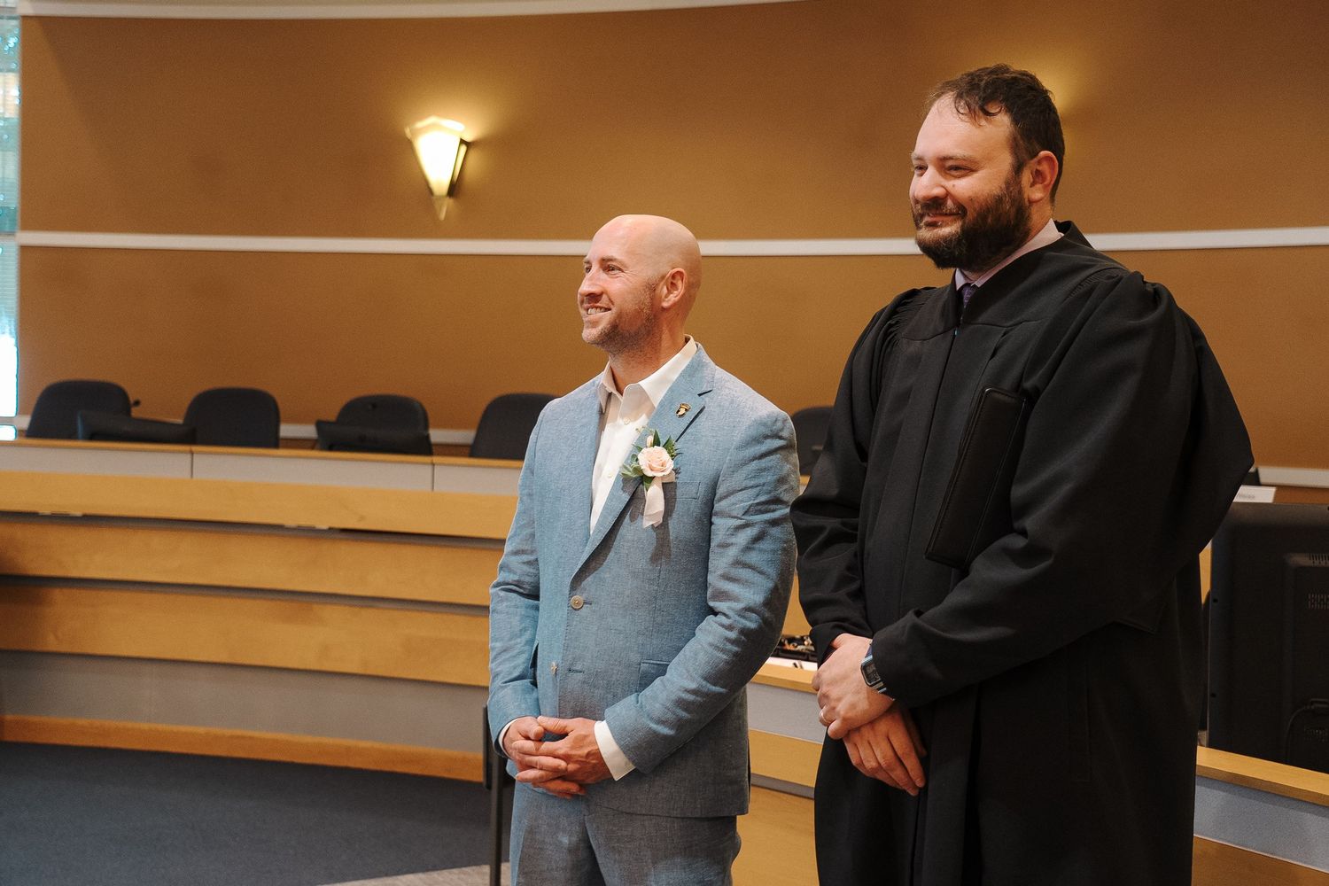 Two people stand in a courtroom setting with one wearing a light blue suit and another in black attire.