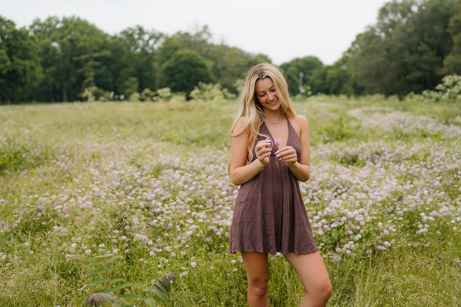 Someone stands in a wildflower meadow wearing a brown sundress while laughing and holding a camera on a sunny day.