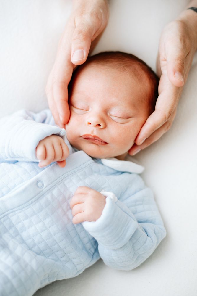 A gently sleeping newborn baby wearing light blue quilted clothing is held by caring hands.