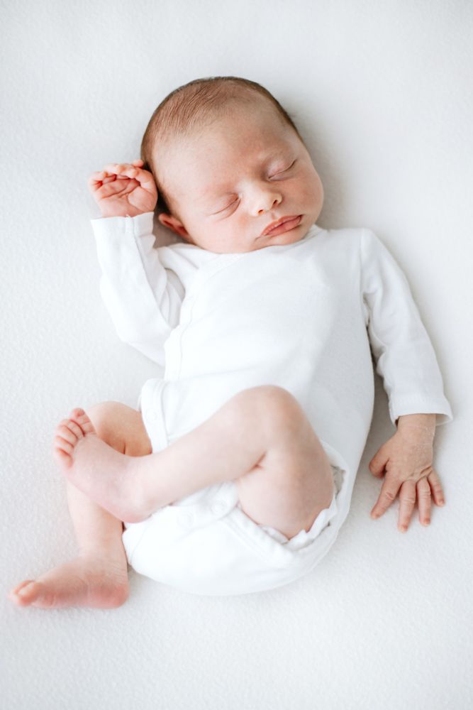 Sleeping newborn baby dressed in white clothing lying on soft white backdrop in artistic photoshoot series.