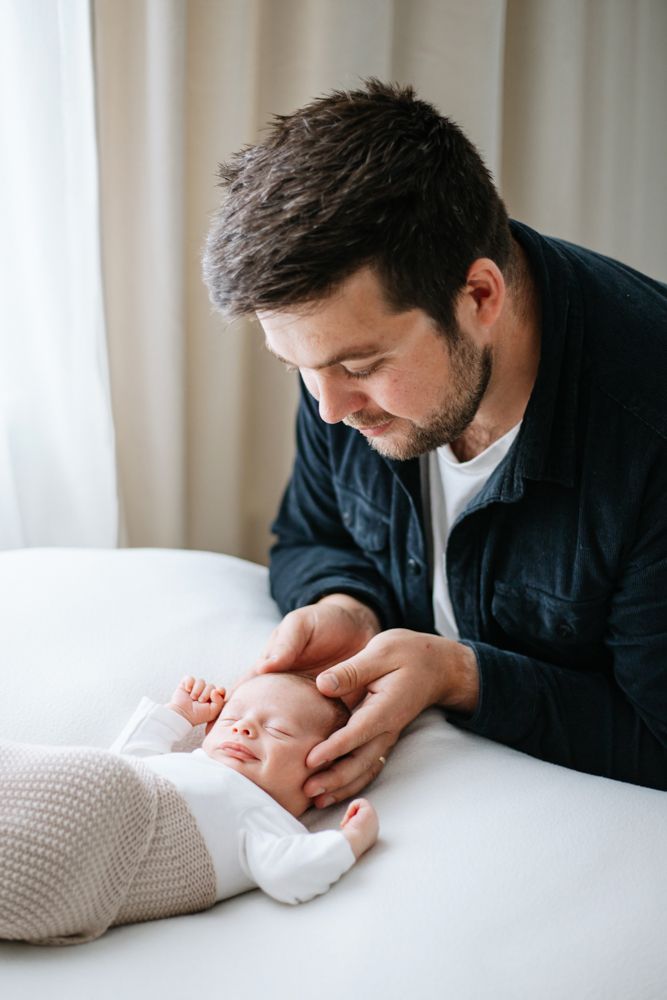 A tender moment as a man in a navy jacket gently touches the head of a sleeping newborn on white bedding.