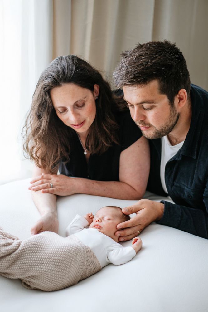 Parents gaze lovingly at their sleeping newborn baby while resting on a white surface with a knit blanket.