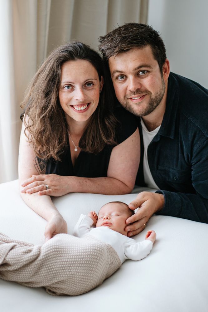 New parents share loving moments with their sleeping newborn baby on white bedding in natural lighting.
