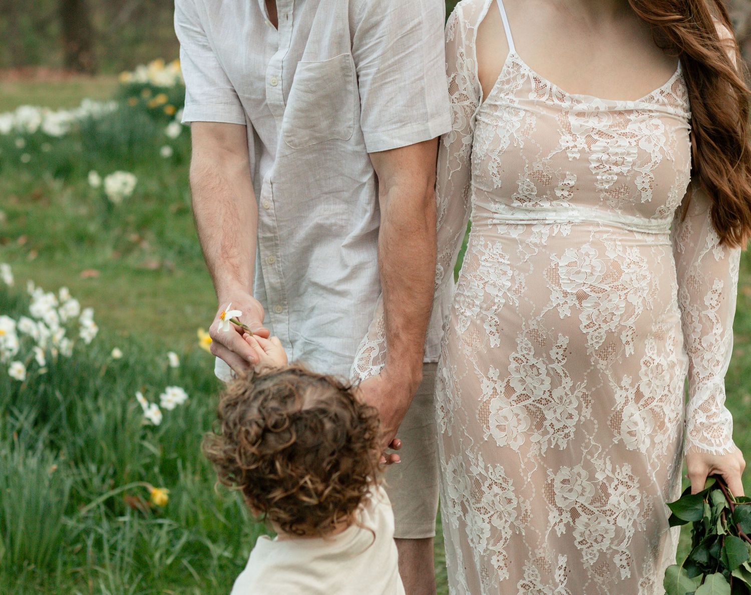 A couple in soft neutral attire holds hands with their toddler while standing in a spring garden with wildflowers.