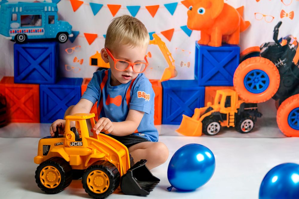 Young child in blue shirt plays with orange construction toy vehicles surrounded by blue balloons and colorful decorations.