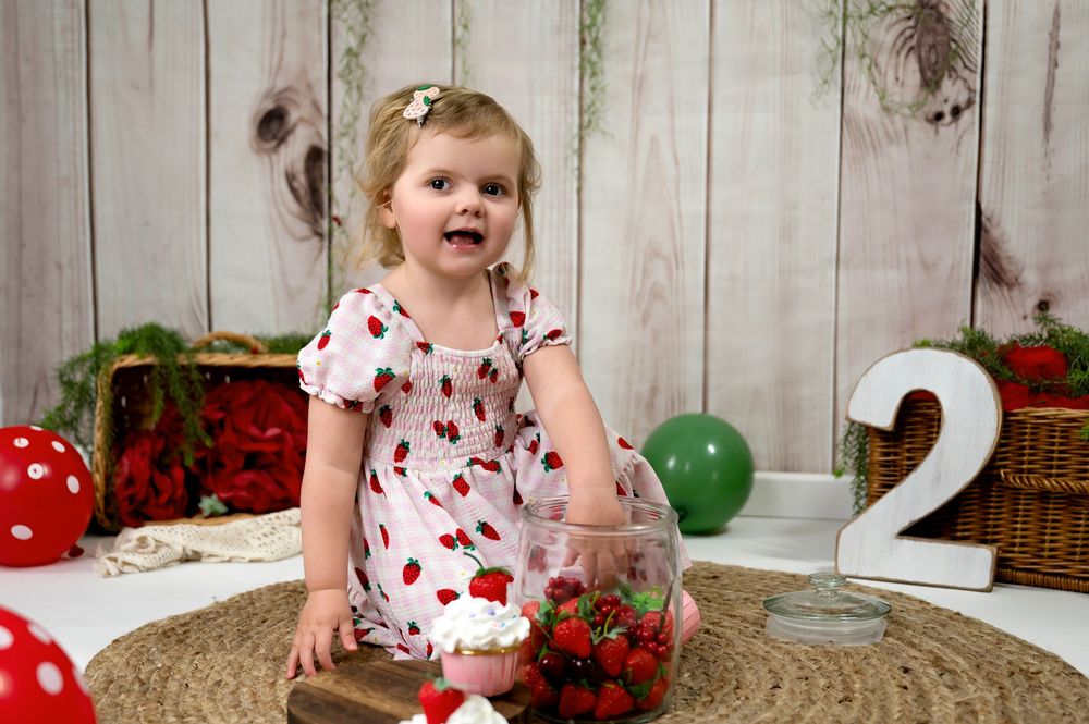 A toddler sits on a round rug with festive decorations including red mushrooms, green balloons and a number 2.