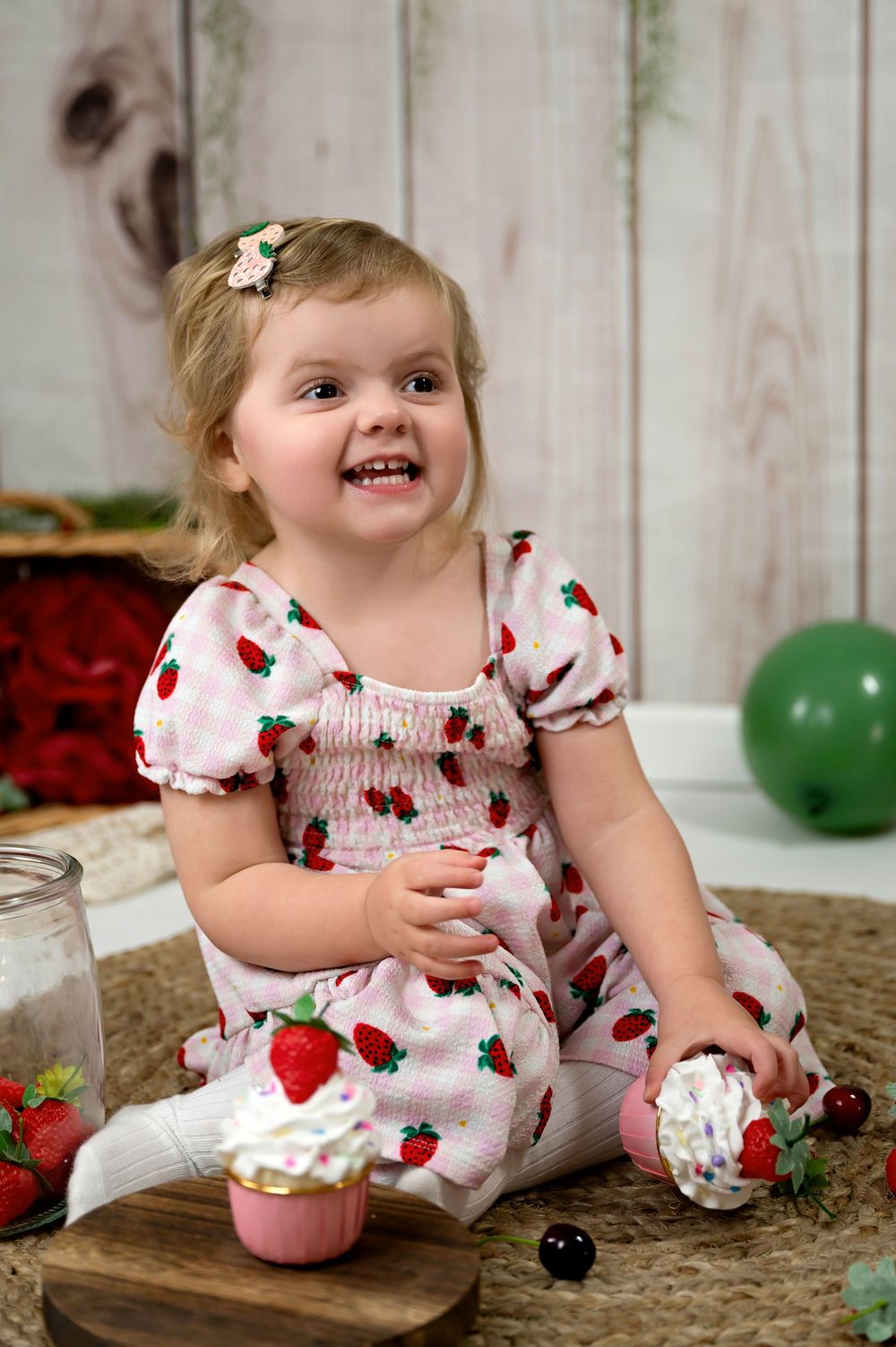 Toddler in white and red dress with cherries pattern sits on rug playing with decorative cupcakes and sweet treats.