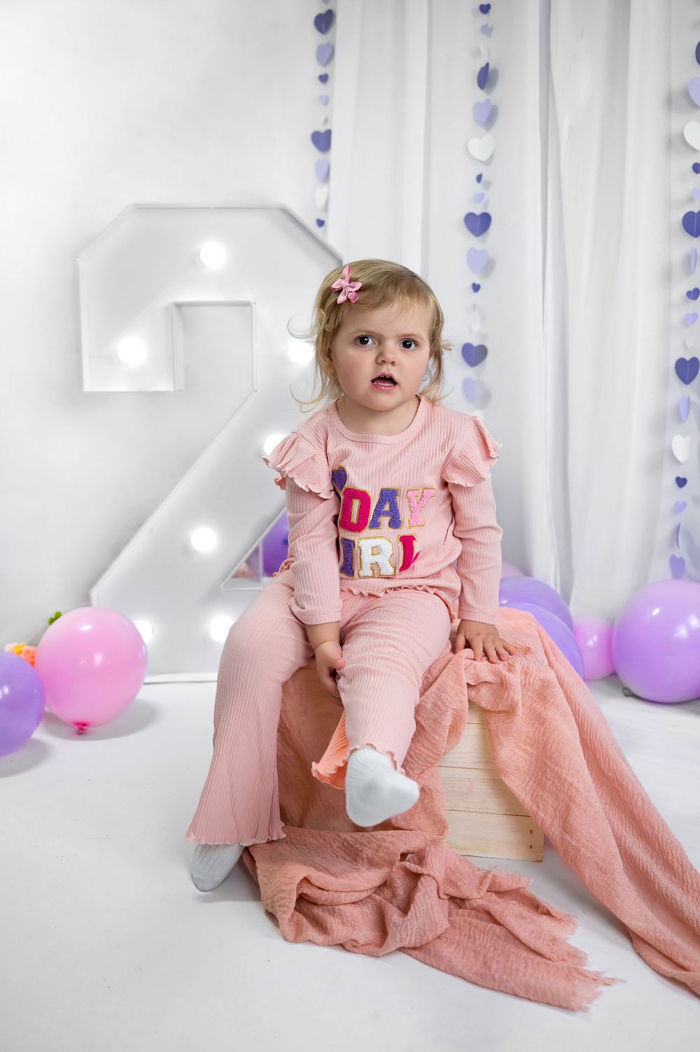 A small child in pink clothing sits on illuminated number 2 with purple and pink balloons and heart decorations.