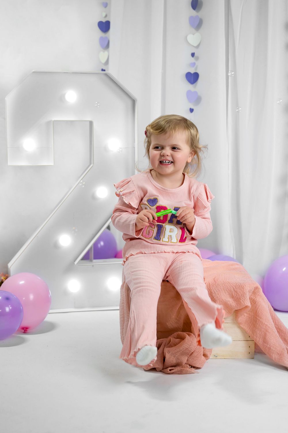 A child in pink outfit sits in front of a large illuminated number 2 with purple and white heart decorations and balloons.