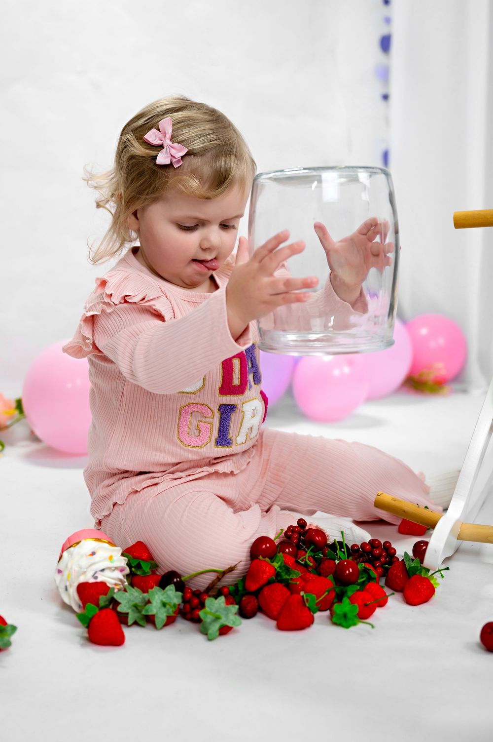Child in pink outfit plays with a glass jar surrounded by strawberries and pink balloons in a bright setting.