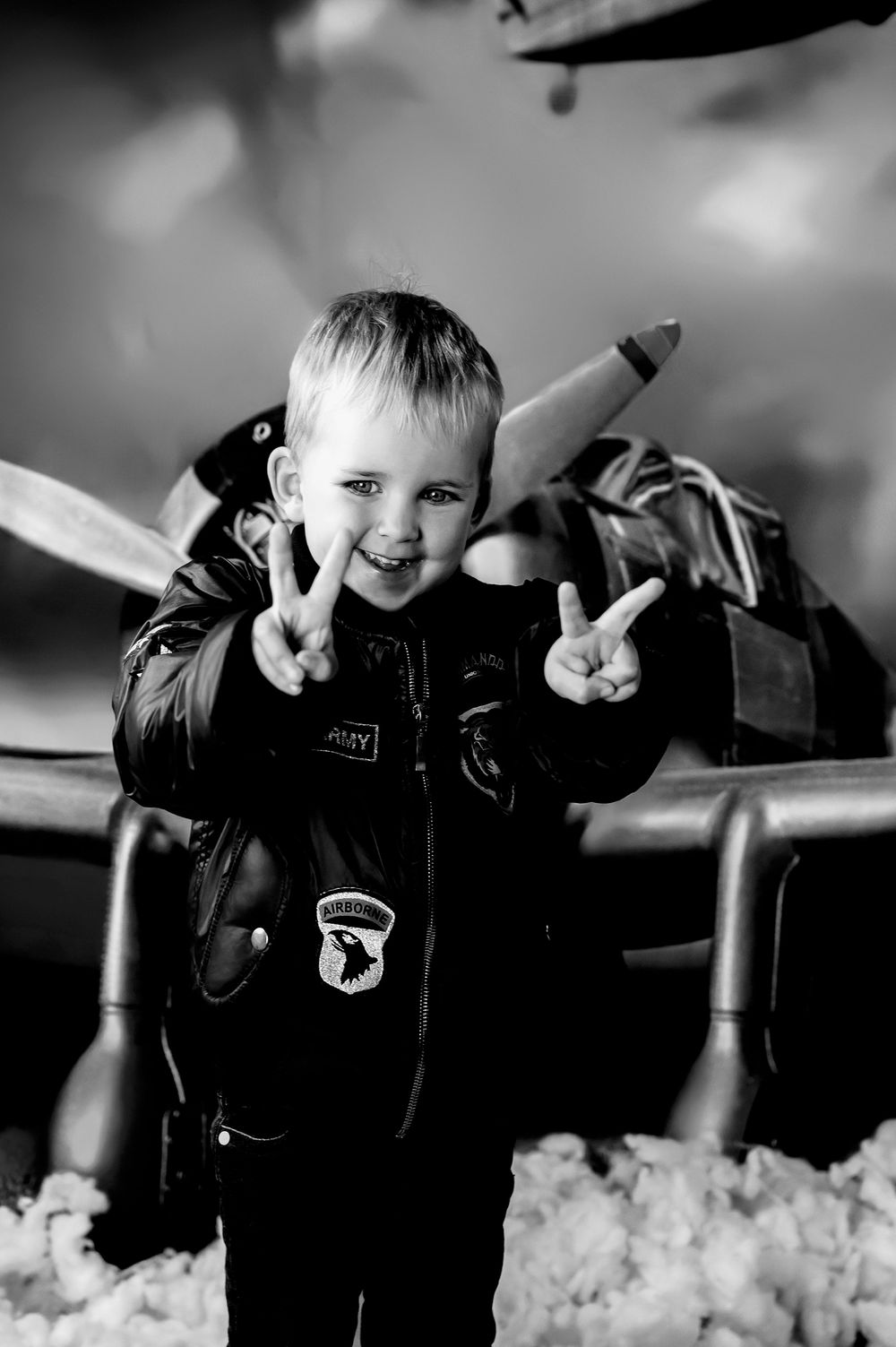 Black and white photo of a young figure in winter clothing making peace signs with both hands while wearing an aviator jacket.