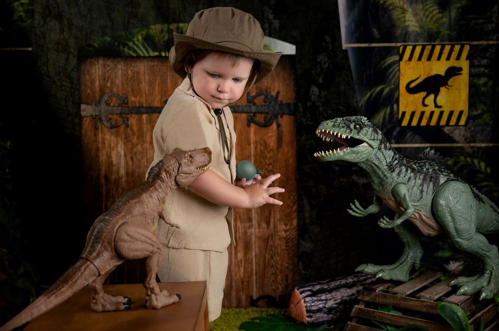 Young child in safari outfit plays with large green dinosaur toy in a themed photo setting with rustic backdrop.
