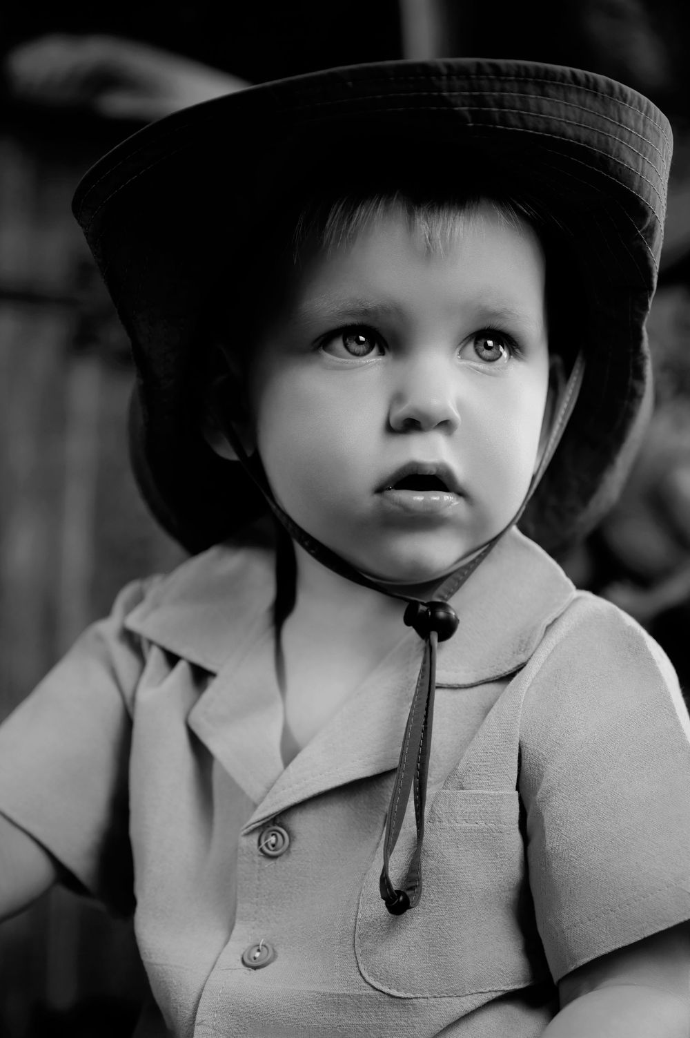 Black and white portrait of small child wearing wide-brimmed hat and button-up coat looking upward with wonder.