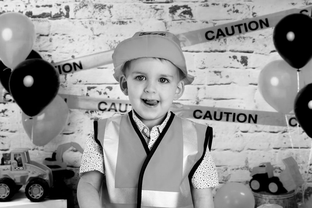 Black and white photo of young child in construction vest and cap with balloons and caution tape party decorations.