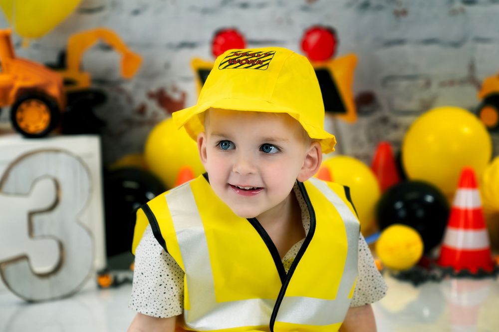 Young child wearing yellow safety vest and hardhat smiles among construction themed party decorations.