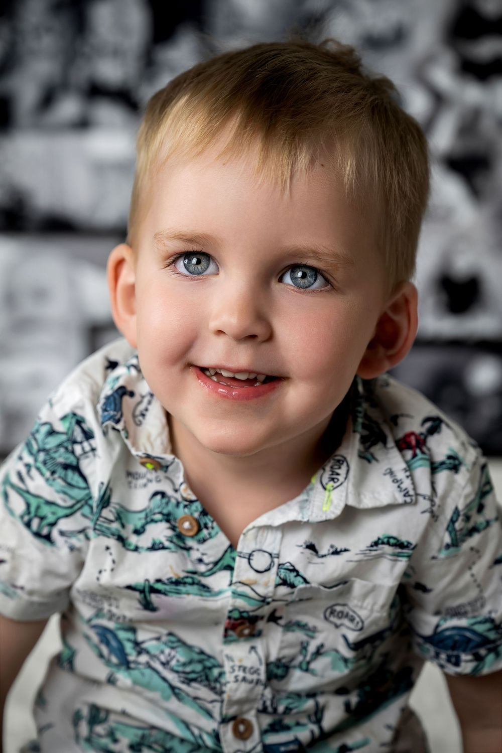 Portrait of a cheerful young child with bright blue eyes wearing a white button-up shirt with dinosaur prints.