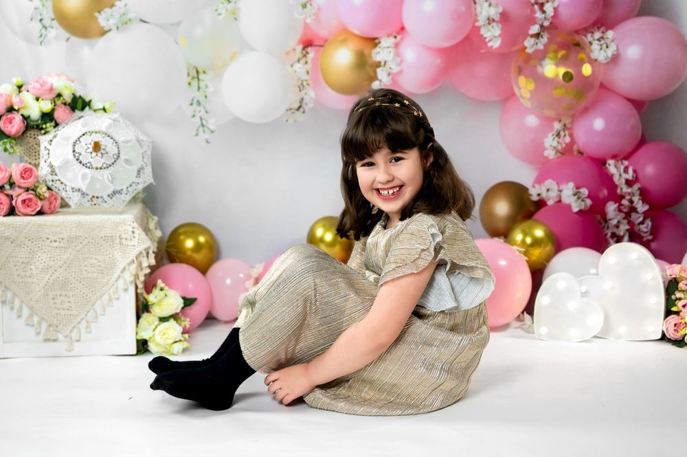 Festive party scene with gold and pink balloon arch, white backdrop and decorative flowers against wall.