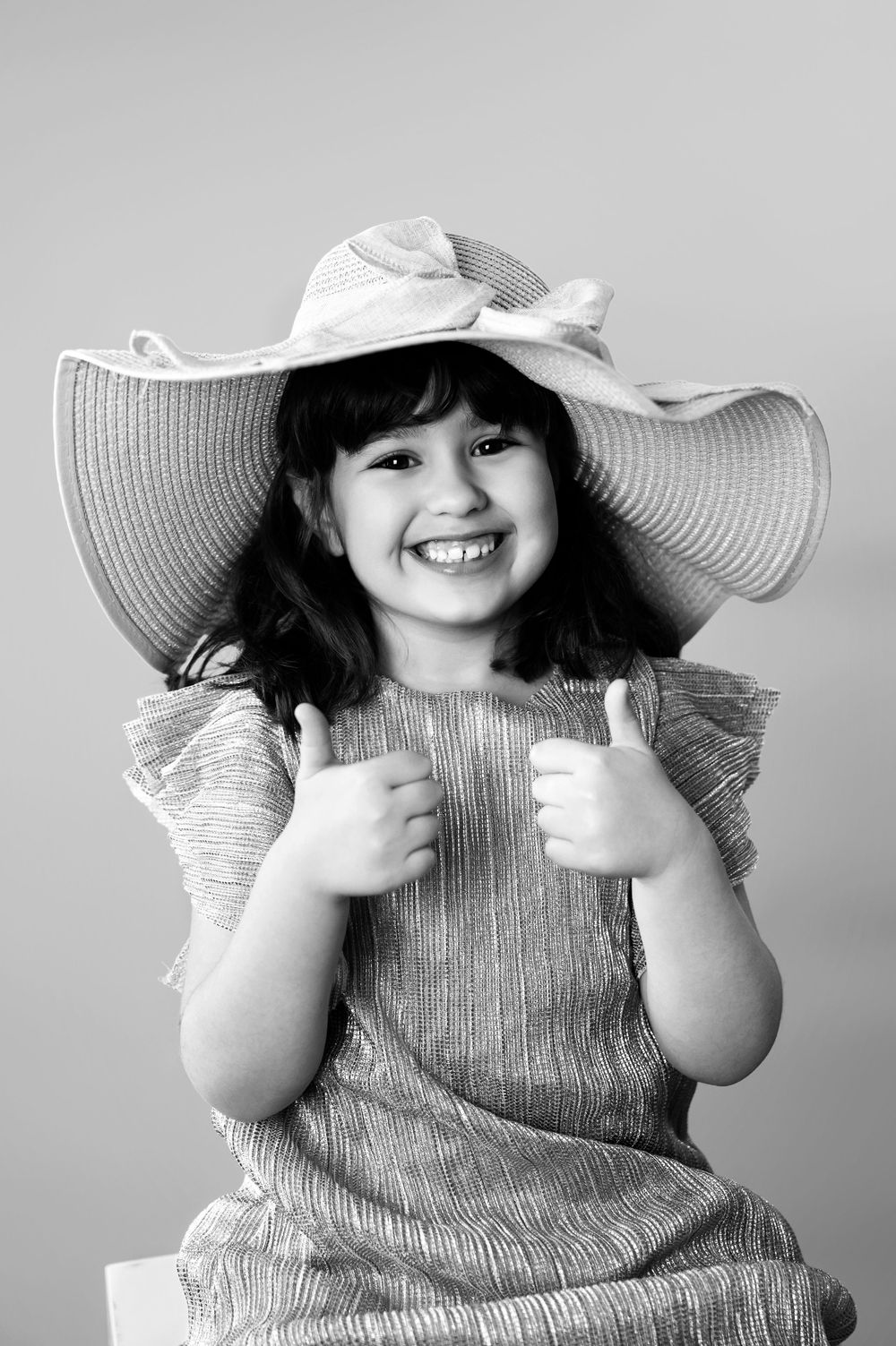 A child wearing a ruffled gray dress and floppy white sunhat poses playfully in a black and white photo series.