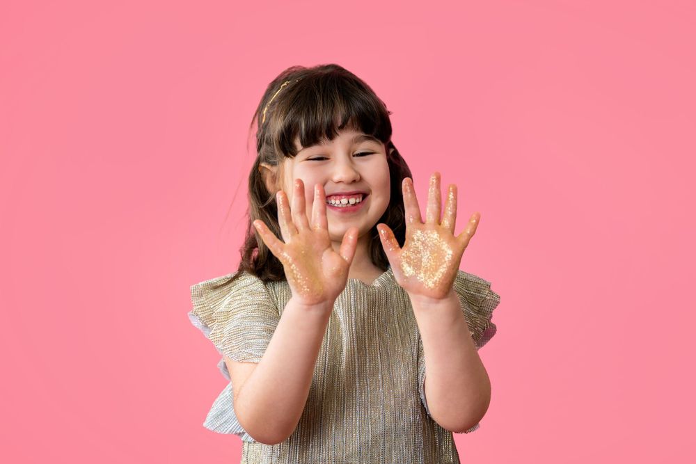 A young person wearing a beige sweater making a playful expression with raised hands against a pink background.